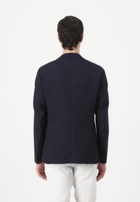 Tengerészkék pamut blézer, amely testre szabott fazonú, éles hajtókájú és egyetlen hátsó szellőzővel rendelkezik, fehér nadrággal párosítva. Gombos mandzsettákkal rendelkezik.