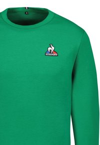 Sudadera verde de cuello redondo con un pequeño logo de Le Coq Sportif en blanco, azul y rojo en el lado izquierdo del pecho, tamaño mediano.