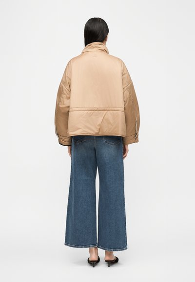 Γυναίκα με μακριά σκούρα μαλλιά που φοράει μπεζ μπουφάν με επένδυση, φαρδιά μπλε τζιν παντελόνια και μαύρα παπούτσια με τακούνι, στέκεται γυρισμένη προς τα πίσω μπροστά σε απλό φόντο.