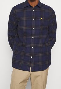 Chemise à carreaux bleu marine avec des accents orange, fabriquée en flanelle. Présente une poche poitrine et des boutons blancs, conçue avec un col classique.