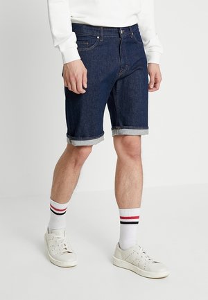 Homme portant un short en denim bleu foncé avec revers, des chaussettes de sport blanches à rayures rouges et noires, et des baskets blanches sur fond blanc.