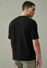 Camicetta nera a maniche corte con texture a coste, vestibilità rilassata e scollatura rotonda, abbinata a pantaloni grigi chiari.
