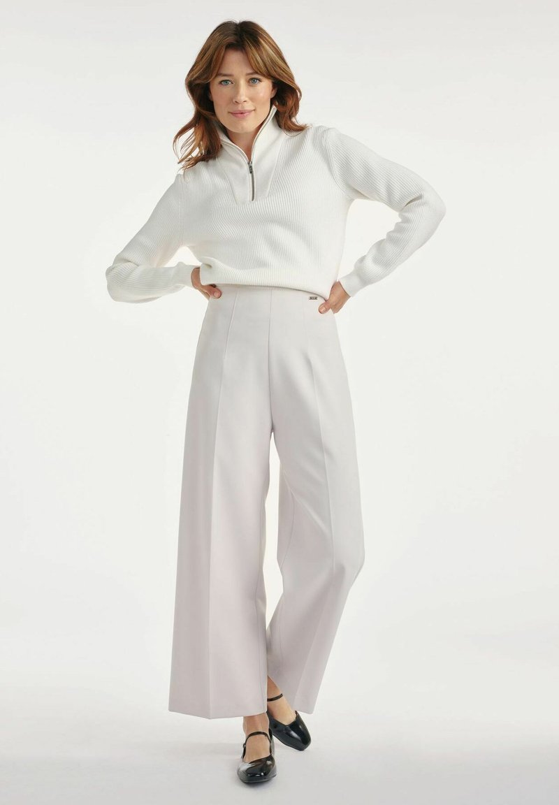 Kobieta z falującymi brązowymi włosami prezentuje biały sweter zapinany na zamek i jasnoszare szerokie spodnie, stojąc pewnie z rękami na biodrach.