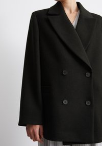 Dvojité černé vlněné kabát s klopovým límcem, velkými černými knoflíky a bočními kapsami. Měkká textura s uvolněným střihem.