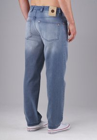 Man wearing light blue relaxed-fit jeans and white Converse sneakers, standing with back facing the camera against neutral background.