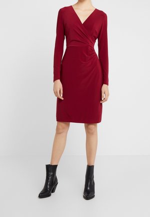 Vrouw draagt een knielange, lange mouwen, dieprode wikkeljurk gecombineerd met zwarte enkellaarzen, staand tegen een effen witte achtergrond.