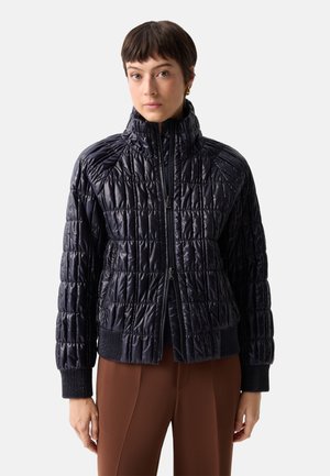Frau trägt eine schwarze gesteppte Jacke mit Reißverschluss und gerippten Bündchen sowie braune Hose, steht vor schlichtem hellem Hintergrund.