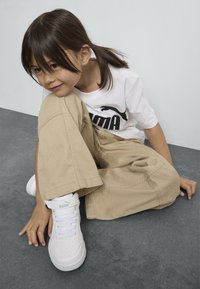 Witte sneakers met een gestructureerde bovenkant en strap, beige wijde broek, en een wit T-shirt met een zwart logo, zittend op een grijs oppervlak.