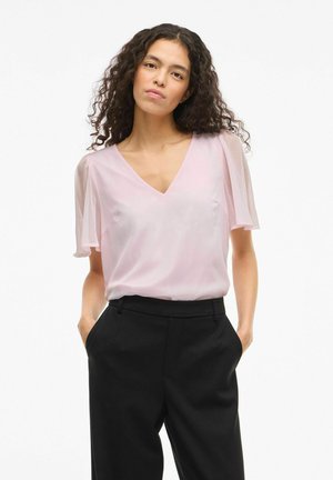 Kobieta o kręconych włosach, ubrana w przezroczystą różową bluzkę z dekoltem w serek oraz czarne spodnie z wysokim stanem, z rękami w kieszeniach, stoi na białym tle.