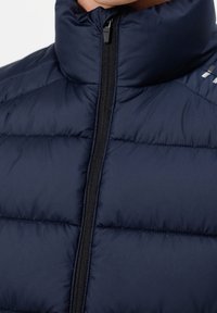 Zbliżenie na osobę ubraną w granatową kurtkę puchową z czarnym zamkiem błyskawicznym z przodu i wysokim kołnierzem.