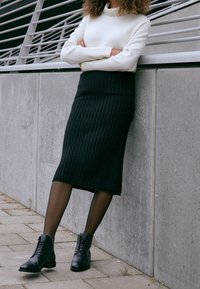 Falda midi de punto acanalado negra combinada con un suéter blanco de manga larga y botas negras de cordones, con un fondo de pared de hormigón.