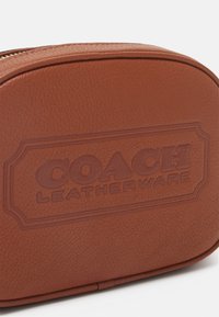 Borsa cosmetica in pelle marrone con logo embossato "COACH LEATHERWARE", forma rotonda e superficie testurizzata. Presenta una chiusura con zip dorata.