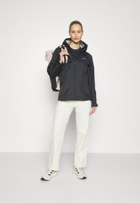 Giacca nera impermeabile con cappuccio, dotata di regolatori, indossata sopra una maglietta nera, abbinata a pantaloni color crema e sneakers.