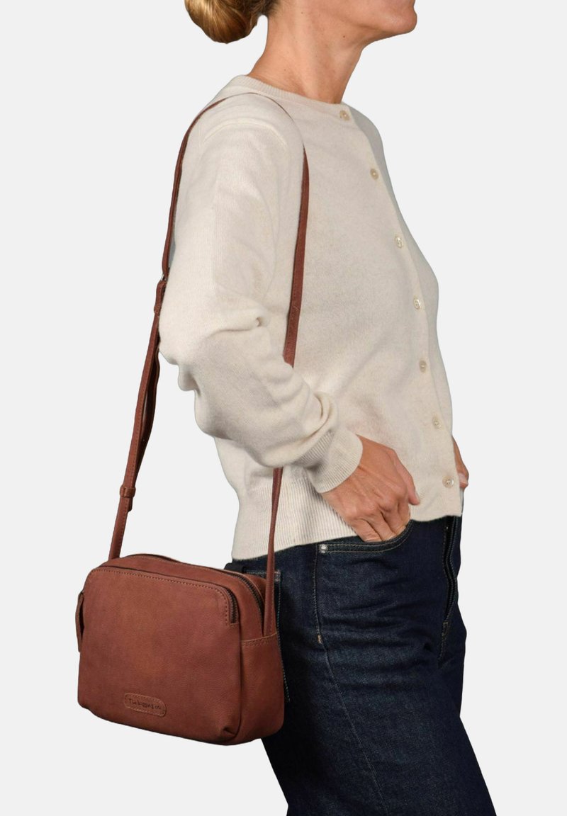Bolso cruzado de cuero marrón con forma rectangular, cierre con cremallera y correa ajustable, llevado sobre un suéter claro y unos jeans oscuros.