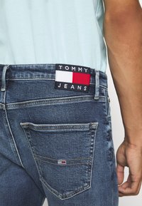 Närbild av person som bär blå denimjeans med Tommy Jeans-märke på midjan och liten logotyp på bakfickan, iklädd en ljusblå skjorta.