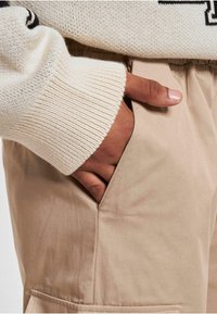 Pantaloni din bumbac bej cu un buzunar lateral, însoțiți de un pulover texturat crem cu manșete cu nervuri și detaliu logo.