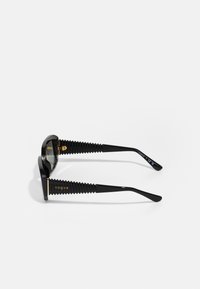 Black sunglasses with a pronounced geometric frame. Textured black temples with ribbed pattern. Gold logo on the side, dark tinted lenses.