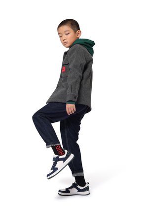 Veste en laine grise avec un patch rouge, couche à capuche verte en dessous, pantalon en denim foncé et baskets bleu marine et blanches avec des semelles en caoutchouc.