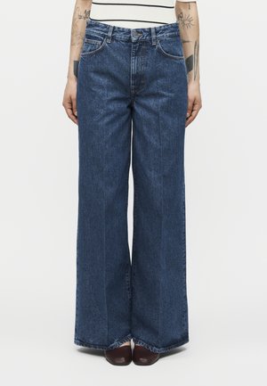 Άτομο που φοράει ψηλόμεσα, φαρδιά σκούρα μπλε τζιν παντελόνια, καφέ παπούτσια και λευκό αμάνικο τοπ με ορατά τατουάζ στα αντιβράχια.