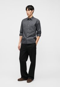 Άνδρας που στέκεται με τα χέρια στις τσέπες, φορώντας σκούρο γκρι πουκάμισο με κουμπιά, μαύρο παντελόνι και καφέ παπούτσια, πάνω σε απλό λευκό φόντο.