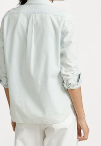 Ljusblå denimskjorta, långa ärmar uppvikta, ryggslits detaljer, avslappnad design, mjuk textur och rundad nederkant.