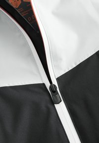 Sort og hvid vindjakke med lynlås foran. Har en tekstureret lynlås og kontrasterende paneldesign. Materialet virker glat.