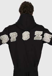 Eine Person mit dunklem, lockigem Haar trägt einen schwarzen Kapuzenpullover mit großen reflektierenden Buchstaben "DPSZE" auf der oberen Rückseite vor einem schlichten Hintergrund.