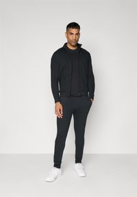 Sweatshirt com fecho preto e calças ajustadas combinando, feitas de tecido macio. Apresenta mangas longas, gola alta e ténis brancos.