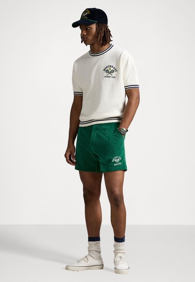 Hvit strikket kortermet skjorte med marineblå stripete mansjetter, parret med grønne kordfløyelsbukser med et logo. Hvite joggesko og en mørk caps.