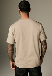 Beige kortærmet t-shirt med rund hals og afslappet pasform, lavet af blødt materiale med en glat tekstur. Sorte denimjeans er synlige.