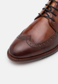 Chaussure oxford en cuir marron avec un bout pointu, des détails de brogue, des lacets noirs et une semelle texturée pour une meilleure adhérence.