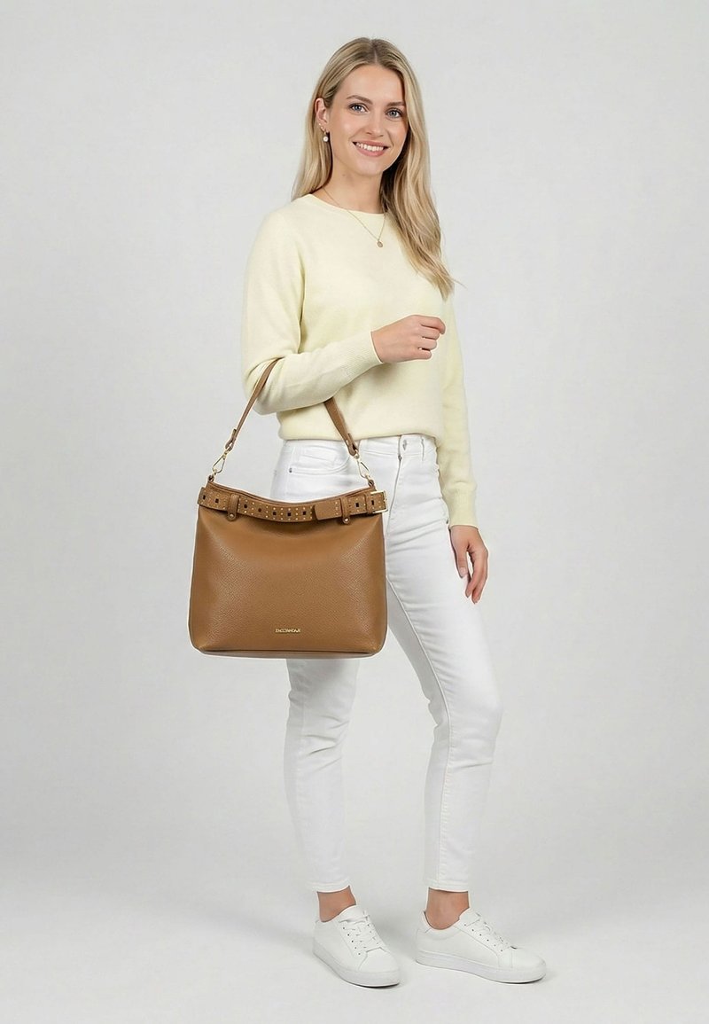 Femme en pull crème et pantalon blanc tenant un grand sac à main en cuir marron avec un détail ceinture, debout devant un fond uni.