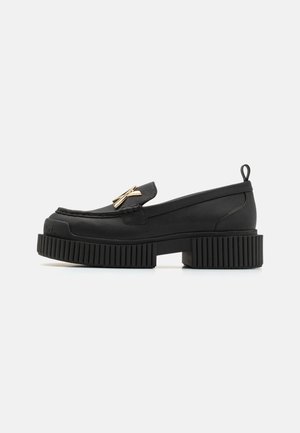 Mocassin en cuir noir avec semelle épaisse à rainures, détail nœud doré sur le cou-de-pied, petit talon carré et languette à l'arrière du col.