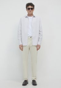 Giovane uomo con occhiali da sole scuri, camicia bianca aperta sopra una maglietta bianca, pantaloni color crema e mocassini marroni scuri, in piedi contro un muro bianco.