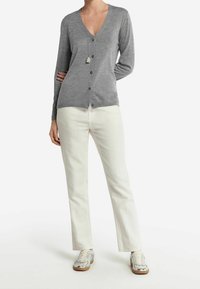 Šedý kardigan s hlubokým výstřihem do V, zapínáním na knoflíky vpředu, dlouhými rukávy a měkkou texturou, v kombinaci s bílými kalhotami a stříbrnými teniskami.