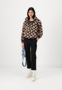 Chaqueta corta negra con flores marrones, pantalones negros, zapatillas blancas y un bolso azul. Modelo de pie contra un fondo blanco.