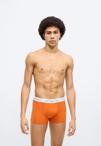 Πορτοκαλί ανδρικά εσώρουχα τύπου μποξέρ με λευκή μέση που φέρει το κείμενο «TOMMY HILFIGER». Το ύφασμα φαίνεται λείο και ελαστικό, σχεδιασμένο για άνεση.