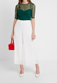 Blusa verde transparente con textura de puntos, combinada con pantalones anchos plisados blancos. Sosteniendo un pequeño bolso rojo; luciendo unos zapatos de tacón puntiagudos blancos.