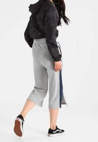 Svart kamouflagemönstrad jacka med huva, grå avklippta sweatpants med sidostripes i marinblått, och svarta sneakers med vita sulor.