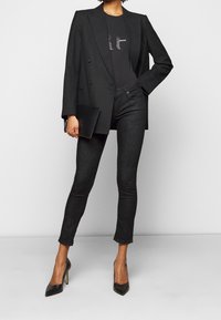 Zwarte double-breasted blazer over een zwart T-shirt met tekst, gecombineerd met donkere skinny jeans en zwarte pumps met spitse neus, terwijl ze een zwarte clutch vasthoudt.