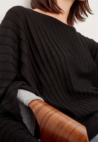 Femme portant un pull côtelé noir à large encolure, assise sur un fauteuil en bois avec une manche gris clair visible en dessous.