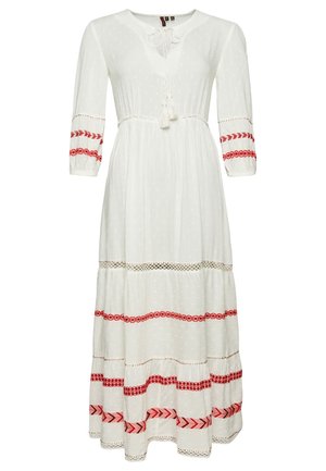 Mujer con cabello rizado que lleva un vestido largo blanco con bordado rojo, sujetando la falda y con zapatillas blancas.