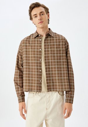 Camisa corta de cuadros marrones con botones en la parte delantera, con cuello puntiagudo y mangas largas, combinada con una camiseta de cuello redondo beige claro debajo.