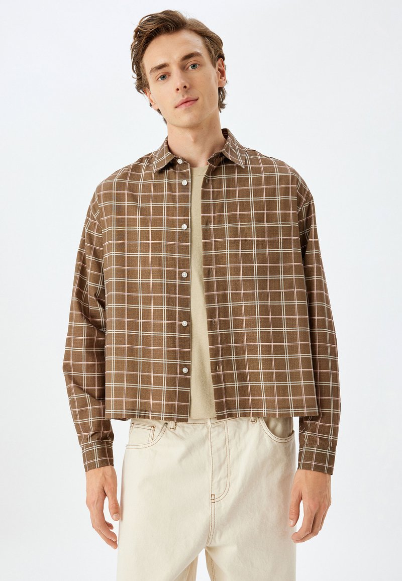 Camisa corta de cuadros marrones con botones en la parte delantera, con cuello puntiagudo y mangas largas, combinada con una camiseta de cuello redondo beige claro debajo.