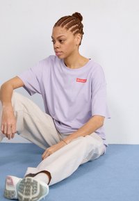 T-shirt viola a maniche corte con logo rosso; abbinato a pantaloni beige larghi e scarpe da ginnastica chiare. Tessuto morbido, design casual.