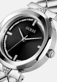 Srebrna nehrđajuća čelična sat sa crnim brojčanikom, dijamantnim detaljima i srebrnim oznakama za sate. Ima okruglo kućište i teksturirani remen.