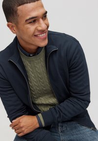 Marinblå zip-up jacka över en grön kabelstickad tröja, kombinerad med mörka jeans. Detaljer inkluderar ribbade ärmmuddar och en skjorta med krage under.