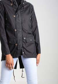 Veste légère noire avec capuche, dotée de boutons sur le devant, poches latérales, cordons de serrage, et une texture lisse et mate.