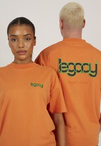 Narančaste pamučne majice s zelenim logotipom: sprijeda imaju mali logotip, dok je na leđima veći logotip s tekstom "Dizajnirano u studiju".