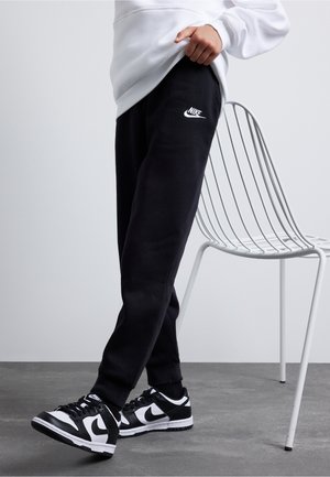 Persona con pantalones deportivos negros de Nike y zapatillas negras y blancas de Nike, de pie junto a una silla metálica blanca sobre un suelo gris.